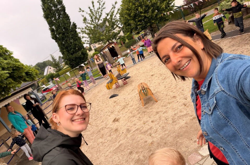 Zwei Frauen posieren für ein Selfie auf einem Sandspielplatz, während im Hintergrund Kinder und Erwachsene spielen. Die Frauen lächeln, während sie die Kamera halten. Im Hintergrund sind Spielgeräte und Bäume zu sehen.