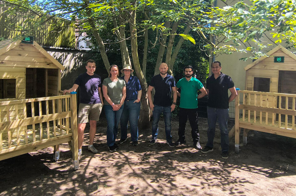 Sechs Personen stehen in einem Garten vor zwei hölzernen Spielhäusern. Sie lächeln und posieren für das Foto. Im Hintergrund sind Bäume und einige grüne Blätter sichtbar. Die Sonne scheint.