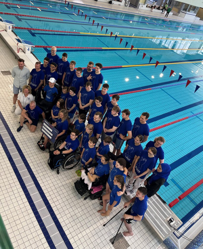 Eine Gruppe von Schwimmern in blauen T-Shirts steht am Rand eines Schwimmbeckens. Es sind etwa 30 Personen, darunter Kinder und Jugendliche. Einige nutzen Rollstühle oder Gehhilfen. Im Hintergrund sind Schwimmbahnen und eine große Glaswand sichtbar. Die Atmosphäre wirkt freundlich und gemeinschaftlich.