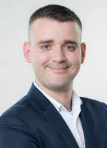 Ein junger Mann mit kurzen, dunklen Haaren und einem leichten Lächeln. Er trägt einen schwarzen Anzug über einem weißen Hemd. Der Hintergrund ist hell und neutral, was den Fokus auf sein freundliches Gesicht legt.