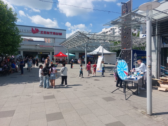 Eine belebte Platzsituation vor einem Einkaufszentrum. Im Hintergrund das Schild "ZENTRUM" und ein Rossmann-Geschäft. Es gibt mehrere Stände, ein Klettergerüst sowie eine Glücksrad-Aktion. Menschen, darunter Kinder und Erwachsene, stehen zusammen, einige unterhalten sich oder warten. Der Himmel ist blau mit wenigen Wolken.
