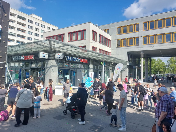 Eine belebte Straße mit vielen Menschen vor einem modernen Gebäude. Im Vordergrund stehen mehrere Personen, darunter Familien mit Kindern und Kinderwagen. Links gibt es eine Kletterwand. An der Fassade des Gebäudes sind die Logos von 'TARGO BANK' und 'PILOT' zu sehen. Im Hintergrund sind weitere Gebäude mit Fenstern in verschiedenen Farben. Der Himmel ist blau mit einigen Wolken.