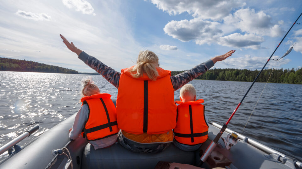 Eine Frau sitzt in einem Boot und hebt die Arme in die Luft. Sie trägt eine orangefarbene Schwimmweste. Neben ihr sitzen zwei Kinder, ebenfalls in orangefarbenen Schwimmwesten. Im Hintergrund ist ein ruhiger See und bewaldete Ufer zu sehen. Der Himmel ist teilweise bewölkt und es reflektiert Sonnenlicht auf dem Wasser.