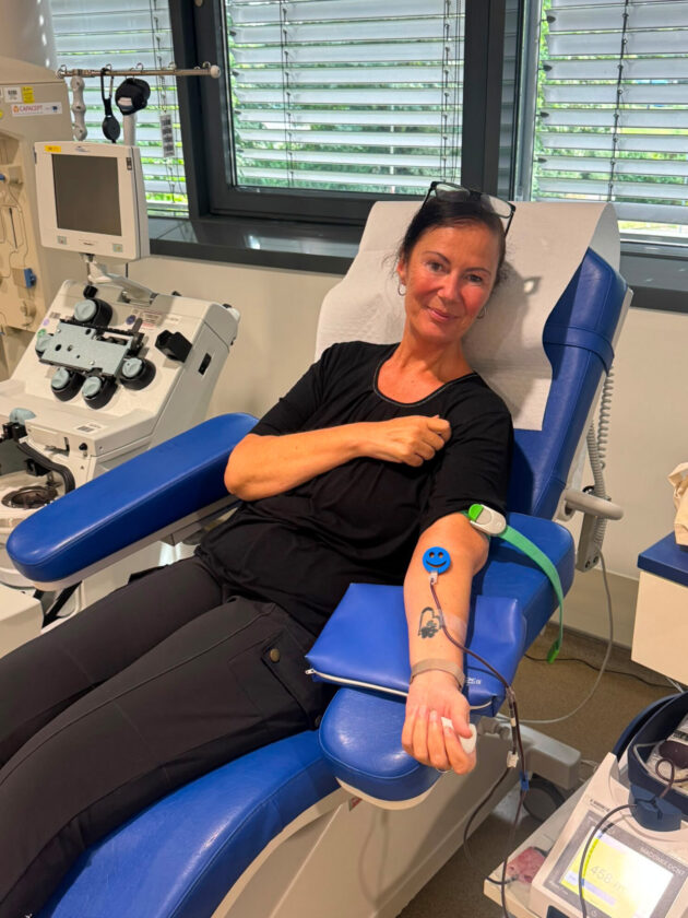 Eine Frau sitzt auf einem blauen Stuhl in einem medizinischen Raum. Sie trägt ein schwarzes Oberteil und hat ihre Arme entspannt. An ihrem linken Arm ist ein IV-Schlauch mit einem lächelnden Aufkleber befestigt. Neben ihr befinden sich medizinische Geräte, darunter ein Monitor und eine Infusionsmaschine, sowie Fenster mit Jalousien.