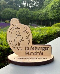Eine Holzskulptur in Form einer stilisierten Familie steht auf einem Tisch. Darauf steht 'Duisburger Bündnis' und '#VorbildlicheVereinbarkeit 2026'.