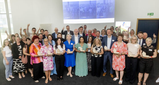 Eine Gruppe von Menschen steht in einem Raum und hält Auszeichnungen in den Händen. Sie lächeln und zeigen Daumen hoch. Im Hintergrund sind weitere Personen zu sehen. Die meisten Teilnehmer tragen formelle Kleidung. An der Wand hängt ein Bildschirm mit Bildern. Der Raum hat große Fenster, durch die Tageslicht strömt.