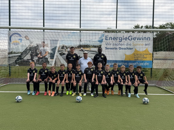 Eine Fußballmannschaft von Kindern posiert auf einem Kunstrasenplatz vor einem Tor. Sie tragen schwarze Trikots und Shorts und haben bunte Fußballschuhe an. Im Hintergrund hängt ein Banner mit dem Text 'EnergieGewinn' und Informationen über Severin Geschier. Es sind insgesamt 14 Kinder und 3 Erwachsene zu sehen, die lächeln und in die Kamera schauen.