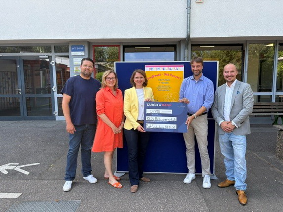 Fünf Personen stehen vor einem Gebäude. Eine Frau in einem gelben Blazer hält einen großen Scheck von der TARGOBANK. Die anderen Personen, darunter ein Mann in einem schwarzen T-Shirt, eine Frau in einem roten Kleid, ein Mann in einem blauen Hemd und ein Mann in einem grauen Anzug, lächeln. Im Hintergrund ist ein Plakat mit der Aufschrift "Support! Das Lernen!" sichtbar.