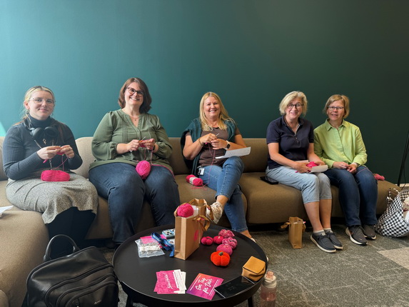 Fünf Frauen sitzen auf einer Couch und stricken mit rosa Wolle. Sie lächeln und unterhalten sich. Vor ihnen steht ein niedriger Tisch mit verschiedenen Handarbeitsutensilien und Getränken. Die Wände sind grün, und der Raum wirkt freundlich und einladend.