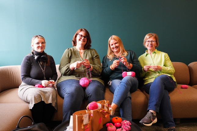 Vier Frauen sitzen auf einem Sofa und stricken mit rosa Wolle. Sie haben unterschiedliche Outfits an und lächeln in die Kamera. Vor ihnen liegen Geschenktüten und bunte Wollknäuel auf einem Tisch.