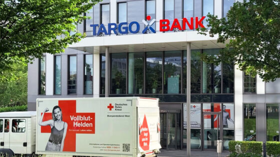 Das Bild zeigt die TARGO BANK, ein modernes Gebäude mit großen Fenstern. Vor dem Eingang steht ein Truck mit der Aufschrift 'Vollblut-Helden', der für Blutspenden wirbt. Auf dem Truck ist ein Bild einer lächelnden Person, die einen Arm hebt. Es sind auch rote und weiße Kreuzsymbole sichtbar, die mit dem Deutschen Roten Kreuz in Verbindung stehen.