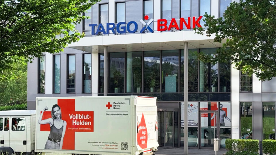 Das Bild zeigt die TARGO BANK, ein modernes Gebäude mit großen Fenstern. Vor dem Eingang steht ein Truck mit der Aufschrift 'Vollblut-Helden', der für Blutspenden wirbt. Auf dem Truck ist ein Bild einer lächelnden Person, die einen Arm hebt. Es sind auch rote und weiße Kreuzsymbole sichtbar, die mit dem Deutschen Roten Kreuz in Verbindung stehen.