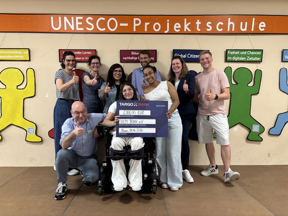 Eine Gruppe von neun Personen steht vor einer Wand mit dem Schriftzug "UNESCO-Projektschule". Im Vordergrund sitzt eine Person im Rollstuhl, die ein großes Scheck-Schild mit der Aufschrift "2.000,00 EUR" von der TARGOBANK hält. Alle Personen zeigen einen Daumen nach oben.