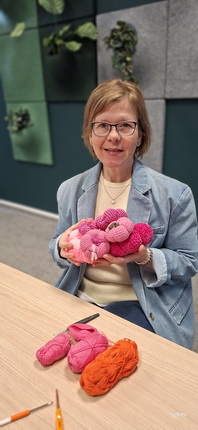 Eine Frau sitzt an einem Tisch und hält mehrere pinkfarbene Wollknäuel in den Händen. Auf dem Tisch liegen weitere Wollknäuel in verschiedenen Pink- und Orangetönen. Im Hintergrund sind grüne Pflanzen und eine graue Wand zu sehen.