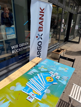 Ein Tisch im Freien mit einem bunten, bemalten Tisch, auf dem mehrere blaue Broschüren der Cargo Bank liegen. Im Hintergrund steht ein Banner mit dem Logo der Cargo Bank. Auf dem Tisch liegen auch einige Informationskarten. Im Fenster des Gebäudes sind Aufkleber sichtbar, und ein Kunde ist hinter dem Fenster zu sehen.