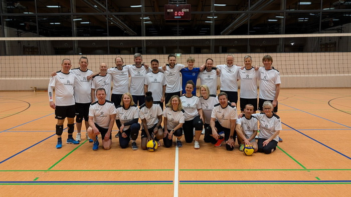 Eine Gruppe von 22 Personen steht in einer Sporthalle vor einem Volleyballnetz. Die meisten tragen weiße T-Shirts mit dunklen Akzenten. Einige halten Volleyballs. Im Hintergrund ist eine Anzeigetafel sichtbar.
