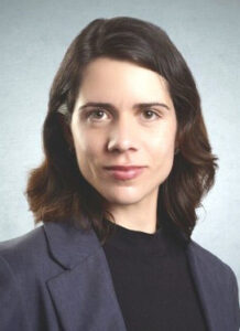 Ein Porträt einer Frau mit mittellangen, lockigen, braunen Haaren, die in einem grauen Blazer und einem schwarzen Oberteil gekleidet ist. Sie blickt direkt in die Kamera mit einem neutralen Gesichtsausdruck. Der Hintergrund ist hell und einfarbig.
