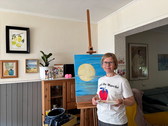 Eine Frau steht in einem Raum vor einer Staffelei mit einem Gemälde, das eine gelbe Sonne auf blauem Hintergrund zeigt. Sie trägt ein T-Shirt mit der Aufschrift "C'est Du Marché" und hält eine Palette in der Hand. Im Hintergrund sind verschiedene Bilder an der Wand und ein Pflanzengefäß sichtbar.