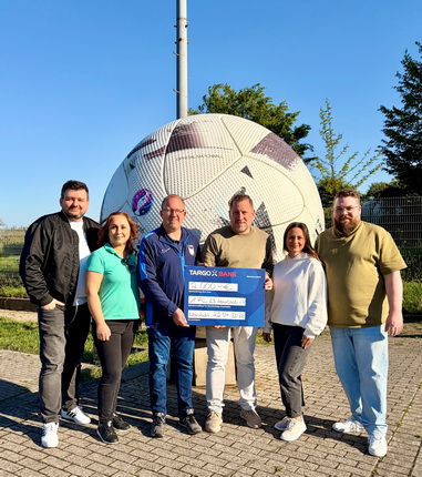 Eine Gruppe von sechs Personen steht vor einem großen Fußball mit einem Banner der TARGOBANK. Die Personen lächeln und posieren für das Foto. Der Fußball ist groß und hat ein auffälliges Design. Im Hintergrund ist eine grüne Landschaft mit Bäumen und einem blauen Himmel zu sehen. Die Gruppe besteht aus Männern und Frauen, die lässige Kleidung tragen.
