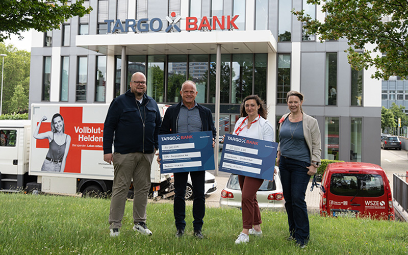 Eine Gruppe von vier Personen steht vor der TARGOBANK. Zwei Männer und zwei Frauen halten große Schecks mit den Beträgen 30.000 € und 100.000 €. Im Hintergrund ist ein Lieferwagen mit der Aufschrift 'Vollblut-Helden' zu sehen.