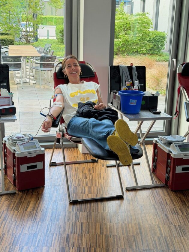 Eine Frau sitzt entspannt in einem Blutspendezentrum und lächelt. Sie trägt ein weißes T-Shirt mit gelber Schrift. Ihr Arm ist verbunden, während sie Blut spendet. Im Hintergrund sind Tische und Stühle in einem grünen Bereich sichtbar.