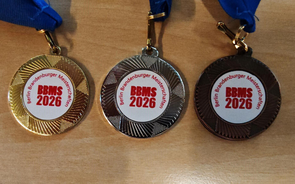 Drei Medaillen liegen auf einem Tisch. Die Medaille links ist goldfarben, die in der Mitte silberfarben und die rechts bronzefarben. Alle Medaillen haben einen weißen Kreis in der Mitte mit rotem Text, der "BBMS 2026" und "Berlin Brandenburger Meisterschaften" anzeigt. Jede Medaille hat ein blaues Band.