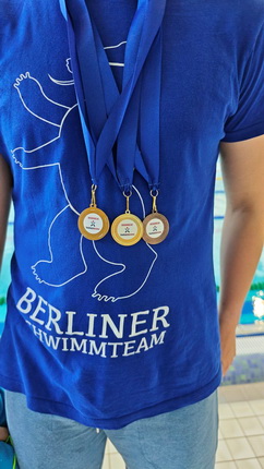 Eine Person trägt ein blaues T-Shirt mit dem Aufdruck "BERLINER SCHWIMMTTEAM" und hat drei Medaillen an einem blauen Band um den Hals hängen. Die Medaillen sind goldfarben mit einem weißen Innenteil.