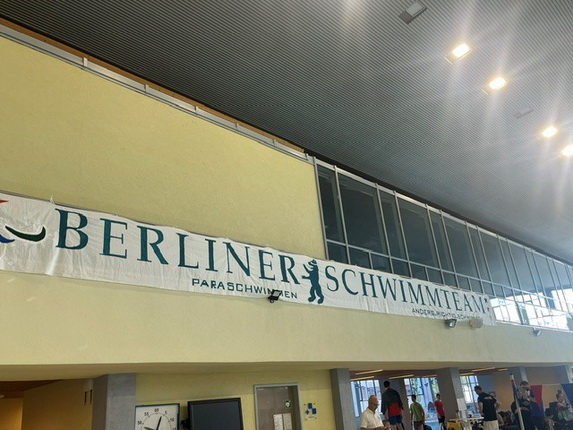 Ein Banner mit dem Text 'BERLINER SCHWIMMTEAM PARA-SCHWIMMEN' hängt in einer Schwimmhalle. Im Hintergrund sind Menschen zu sehen, die sich in einem offenen Raum bewegen.