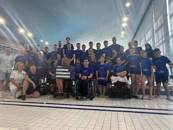 Eine Gruppe von jungen Schwimmern und Betreuern steht in blauen T-Shirts am Beckenrand eines Schwimmbades. Einige Teilnehmer sitzen im Rollstuhl. In der Mitte hält jemand ein Schild hoch. Die Atmosphäre ist fröhlich und gemeinschaftlich.