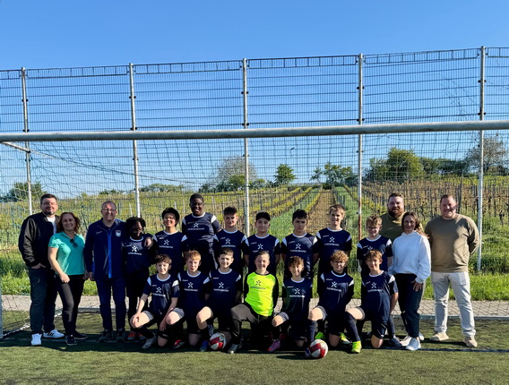 Eine Gruppe von Fußballspielern und Trainern steht im Freien auf einem Fußballfeld. Die Spieler tragen dunkelblaue Trikots mit einem weißen Logo und schwarze Shorts. In der Mitte steht ein Torwart in einem grünen Trikot. Im Hintergrund ist ein grünes Feld und ein Zaun sichtbar. Die Gruppe lächelt und posiert für das Foto, das bei sonnigem Wetter aufgenommen wurde.