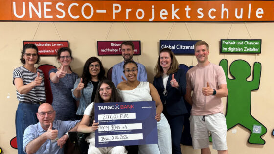 Eine Gruppe von zehn Personen steht vor einer Wand mit dem Schriftzug "UNESCO-Projektschule". Sie zeigen alle den Daumen nach oben. Im Vordergrund hält eine Person einen großen Scheck mit dem Betrag von 2.000,00 EUR, der an die Organisation VKM Bonn e.V. gerichtet ist. Das Datum auf dem Scheck ist der 01.06.2026. Hintergrundinformationen sind in verschiedenen Farben an der Wand sichtbar.