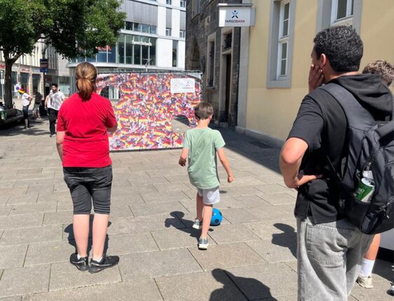 Auf dem Bild sind mehrere Personen auf einer Straße zu sehen. Eine Frau mit langen Haaren trägt ein rotes T-Shirt und schwarze Shorts. Ein Junge in einem grünen T-Shirt und weißen Shorts läuft mit einem Fußball. Ein Mann mit dunklen Haaren steht mit einer Hand am Kinn, während er in die Richtung des Jungen schaut. Im Hintergrund steht ein buntes Kunstwerk oder eine Wand, die mit verschiedenen Farben und Mustern geschmückt ist, und einige Menschen gehen vorbei. Es ist ein sonniger Tag.
