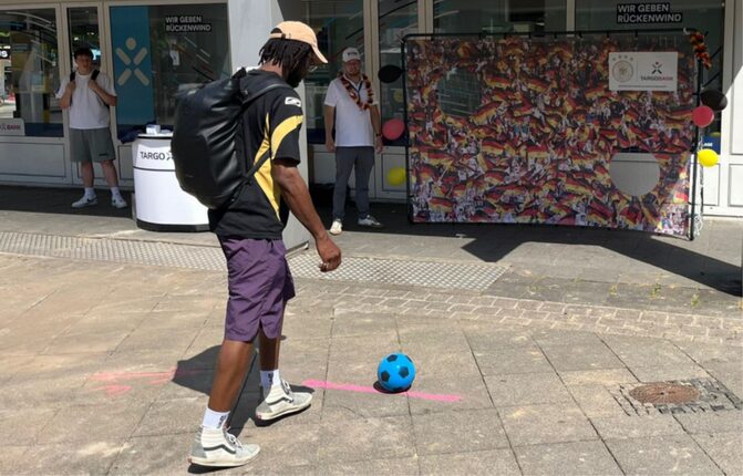 Ein junger Mann mit schwarzem T-Shirt und Rucksack steht auf einem Platz und bereitet sich darauf vor, einen blauen Fußball zu treten. Im Hintergrund ist eine bunte Wand mit einer Sammlung von Bildern, die die Farben der deutschen Flagge zeigt. Weitere Personen stehen hinter ihm, die an einer Veranstaltung teilnehmen. Es ist sonnig und hell.