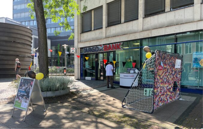 Die TARGOBANK-Filiale hat eine bunte Dekoration mit Fähnchen. Vor der Bank steht ein Fußballtor mit buntem Netz. Zwei Personen sind sichtbar, eine davon trägt ein weißes T-Shirt. Es gibt ein Werbeplakat für die Bank in der Nähe.
