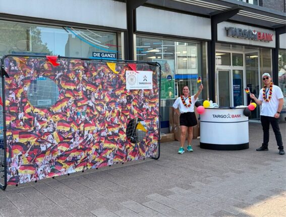 Vor einem TARGOBANK-Geschäft sind zwei Personen zu sehen, die an einer Promotion teilnehmen. Eine Frau trägt eine weiße Bluse und schwarze Shorts, während sie mit einer kleinen Fahne winkt. Der Mann neben ihr trägt eine Sonnenbrille und steht mit einer Wasserpistole. Im Hintergrund hängt ein großes Banner mit vielen deutschen Flaggen und Löchern zum Durchschießen. Ein Stehtisch mit dem TARGOBANK-Logo ist ebenfalls sichtbar.