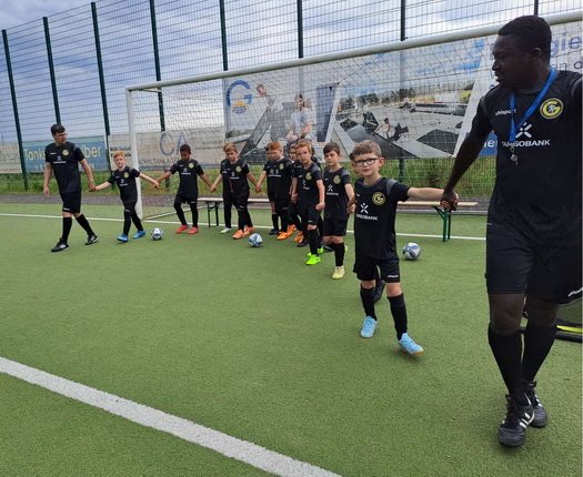 Eine Gruppe von Kindern in schwarzen Fußballtrikots steht auf einem Kunstrasenplatz in einer Reihe. Sie halten sich an den Händen und schauen in die Richtung eines Trainers, der ihnen Anweisungen gibt. Im Hintergrund sind zwei Tore und einige Fußbälle zu sehen. Es ist bewölkt und die Umgebung wirkt sportlich.