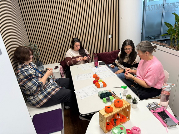 Eine Gruppe von vier Frauen sitzt an einem Tisch in einem modernen Raum mit Holzverkleidung. Sie arbeiten an einem Häkelprojekt, umgeben von Garn und Anleitungen. Auf dem Tisch liegen bereits gehäkelte orangefarbene Kürbisse. Die Frauen scheinen konzentriert und freundlich zu sein.