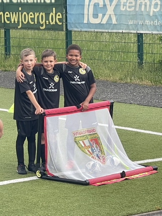 Drei Kinder stehen auf einem Fußballfeld und posieren für ein Foto. Sie tragen schwarze Fußballtrikots mit einem Logo. Zwischen ihnen steht ein Fußballnetz, auf dem ein Wappen sichtbar ist. Im Hintergrund sind Werbebanner und ein grüner Zaun zu sehen.