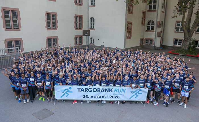 Eine große Gruppe von Teilnehmern des TARGOBANK RUN in Duisburg versammelt sich vor einem Gebäude. Alle tragen blaue T-Shirts und heben die Hände, um zu posieren. Ein Banner mit der Aufschrift 'TARGOBANK RUN 26. AUGUST 2026' ist gut sichtbar. Die Atmosphäre ist fröhlich und energetisch.