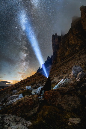 Eine Person sitzt auf einem felsigen Gelände unter einem klaren Nachthimmel. Ein kräftiger Lichtstrahl aus einer Taschenlampe leuchtet in den Himmel, wo die Milchstraße sichtbar ist. Im Hintergrund sind hohe, steile Felsen zu sehen, die die Landschaft umrahmen. Die Szenerie vermittelt ein Gefühl von Weite und Ruhe in der Natur.