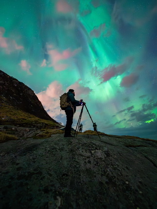 Ein Fotograf steht auf einem Felsen und richtet seine Kamera auf die Nordlichter, die den Himmel in lebhaften Grüntönen erleuchten. Die Landschaft ist bergig und die Wolken sind leicht sichtbar. Im Hintergrund sind Silhouetten weiterer Fotografen zu erkennen.