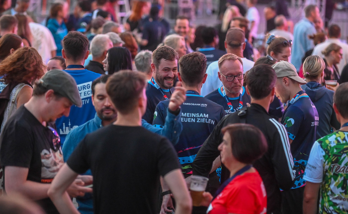 Eine lebhafte Menschenmenge bei einem Event, viele tragen T-Shirts mit Sponsorenlogos. Einige Personen lächeln und unterhalten sich. Der Hintergrund zeigt eine bunte Beleuchtung.