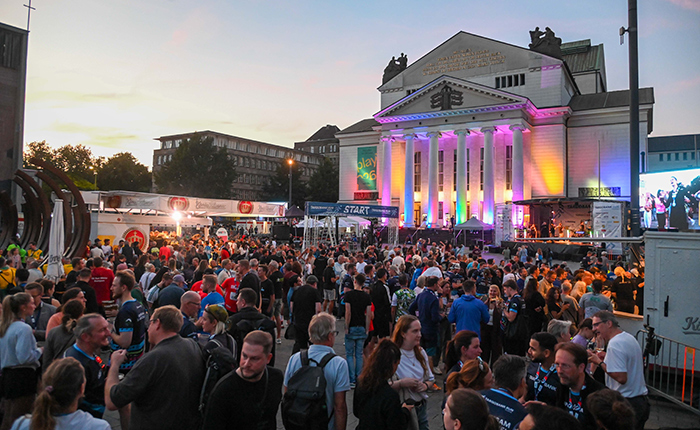 Eine große Menschenmenge versammelt sich vor einem historischen Gebäude mit Säulen, das abends beleuchtet ist. Verkaufsstände und Bühnen sind sichtbar, während die Leute miteinander interagieren und das Event genießen. Der Himmel ist in sanften Farben der Dämmerung gehalten.