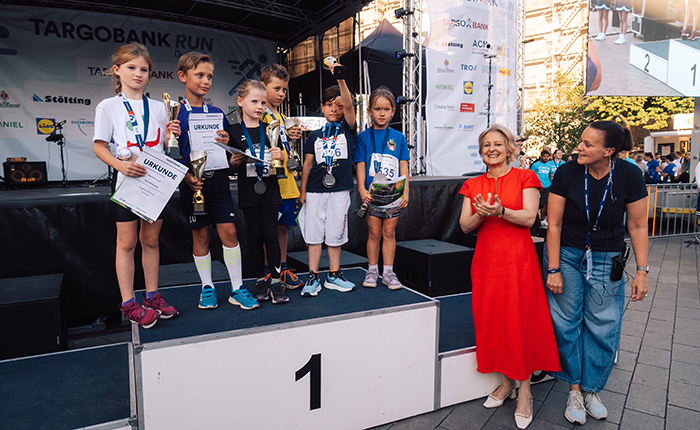 Eine Gruppe von Kindern steht auf dem ersten Platz einer Bühne beim TARGOBANK RUN. Sie halten Medaillen und Pokale. Eine Frau in einem roten Kleid applaudiert, während eine andere Frau in Jeans und T-Shirt lächelt.