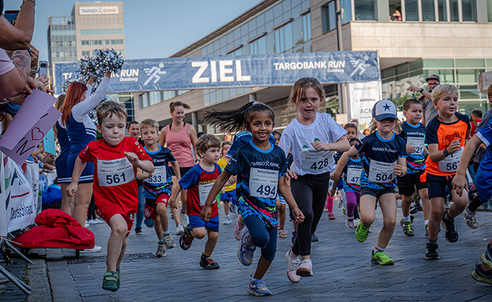 Eine Gruppe von Kindern läuft energisch in einem Rennen, das Teil des TARGOBANK RUN ist. Einige tragen nummerierte Shirts und lächeln, während sie die Ziellinie überqueren. Im Hintergrund ist ein Banner mit der Aufschrift 'ZIEL' zu sehen.