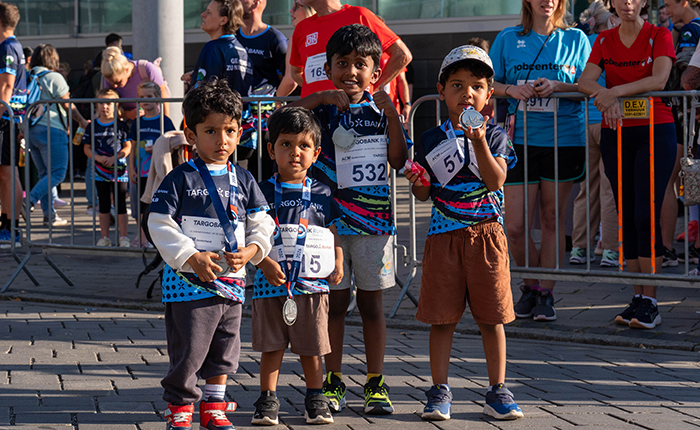 Vier Kinder stehen in sportlicher Kleidung mit Medaillen am Hals. Sie sind in einer Menschenmenge bei einer Veranstaltung, umgeben von anderen Teilnehmern und Zuschauern. Die Kinder lächeln, während sie in die Kamera schauen. Im Hintergrund sind weitere Teilnehmer und Zuschauer zu sehen, die sich auf das Event vorbereiten.