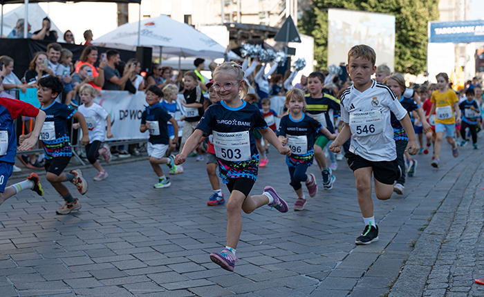 Eine Gruppe von Kindern läuft beim Rennen. Die Kinder tragen sportliche Kleidung und Nummern auf ihren Shirts. Einige Zuschauer sind im Hintergrund zu sehen, die anfeuern. Die Atmosphäre ist lebhaft und die Straße ist mit Steinen gepflastert.