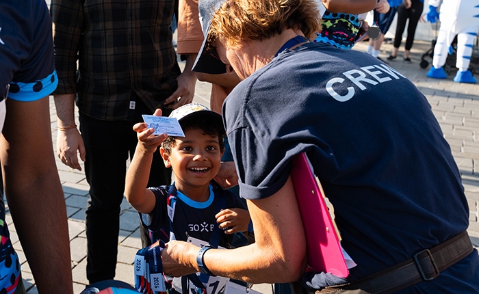 Ein lächelndes Kind hält stolz eine Karte in der Hand, während eine Mitarbeiterin mit einem blauen T-Shirt und der Aufschrift 'CREW' ihm ein Armband anlegt. Im Hintergrund sind andere Teilnehmer und Zuschauer zu sehen, die sich an einem Veranstaltungsort versammeln.