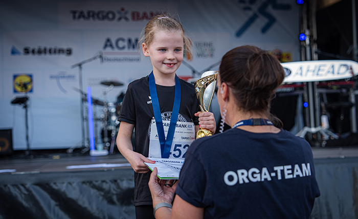 Ein Mädchen mit einem Medaille und einer Trophäe steht auf einer Bühne und erhält eine Urkunde von einer Frau im ORGA-TEAM-Shirt. Im Hintergrund sind Banner und eine Band sichtbar.