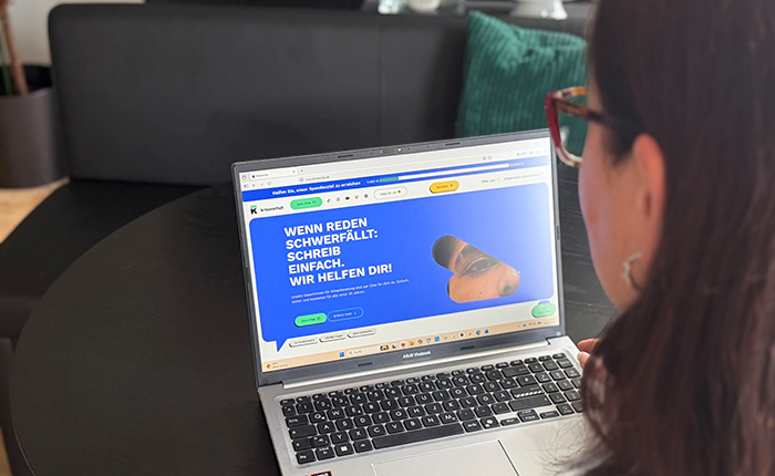 Eine Person sitzt vor einem Laptop, auf dem eine Webseite mit dem Text 'WENN REDEN SCHWERFÄLLT: SCHREIBE EINFACH. WIR HELFEN DIR!' sichtbar ist. Im Hintergrund ist ein dunkles Sofa und ein grünes Kissen zu sehen.